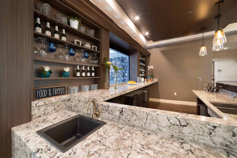 A modern kitchen or bar area with marble countertops, a black sink with a bronze faucet, and wooden shelves holding glasses, bottles, and decorative items. There are pendant lights hanging from the ceiling and a sign on the counter that reads 'FOOD & FRIENDS'.