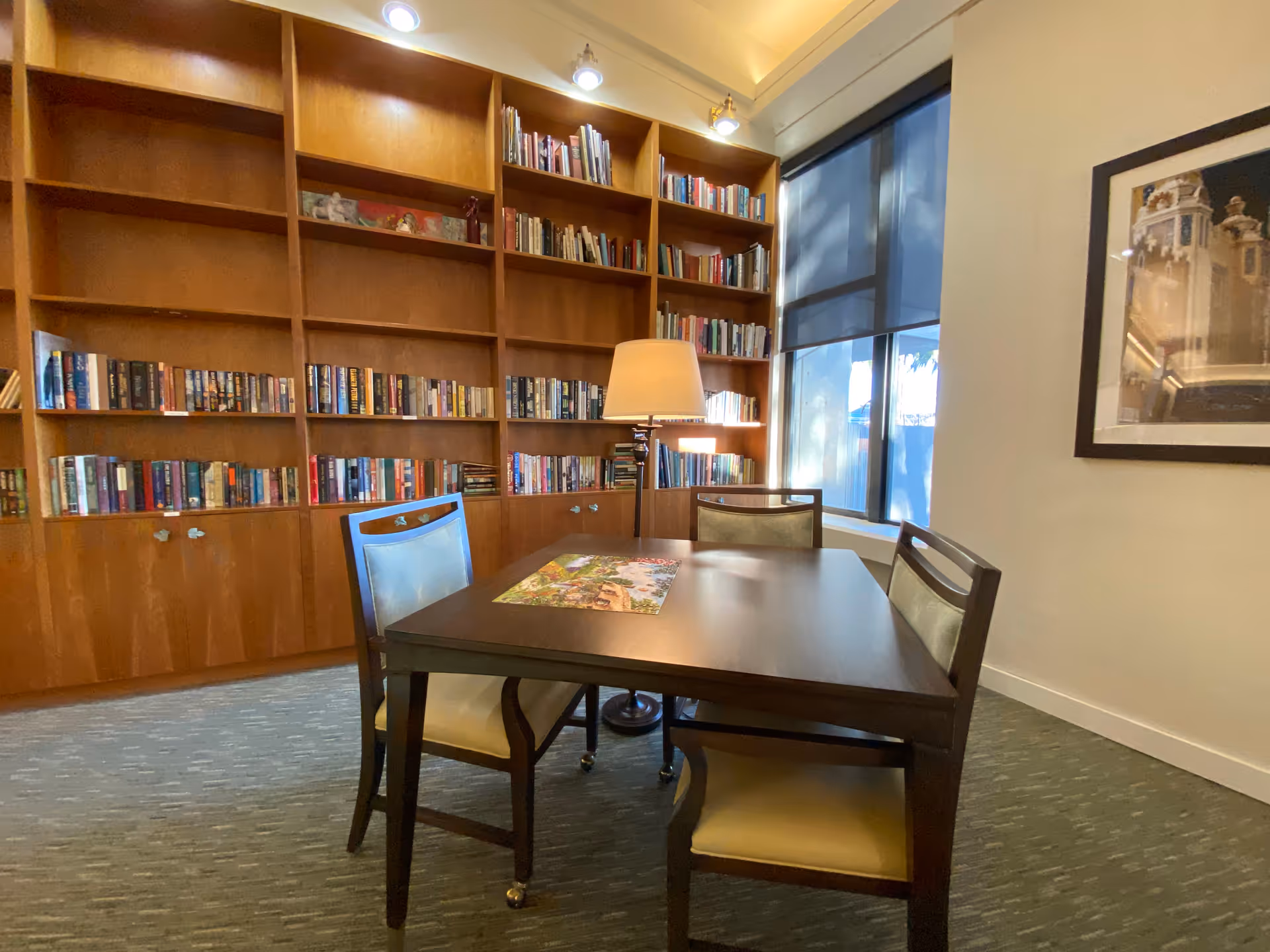 A cozy reading room with a wooden table and four chairs, a floor lamp, large wooden bookshelves filled with books, a window with a blue shade, and a framed picture on the wall.