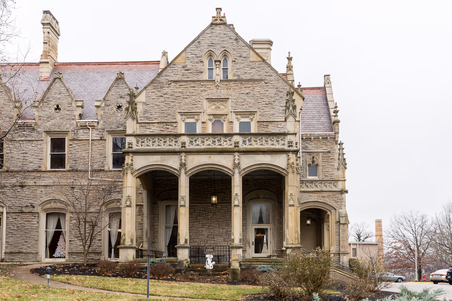 Front exterior view of a large historic stone building with ornate architectural details, arched porch, and multiple windows. The building is surrounded by a lawn with some shrubs and trees, and there are cars visible in the background.