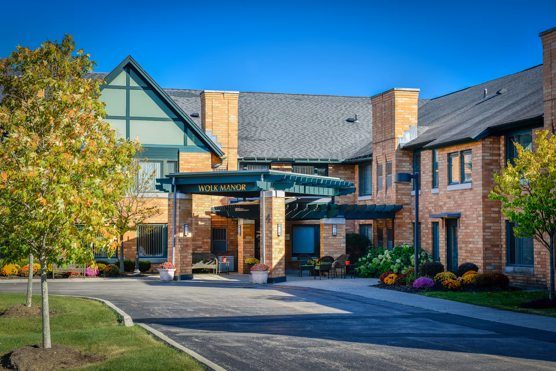 The front entrance of Wolk Manor Assisted Living, a two-story brick building with a covered entry, landscaping, and outdoor seating.
