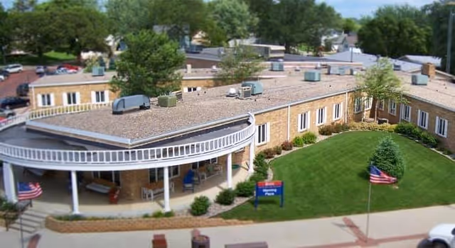 Exterior view of a single-story senior living facility building with a curved covered porch supported by white columns. The building is surrounded by a well-maintained lawn with small bushes and trees, and two American flags are visible near the sidewalk.