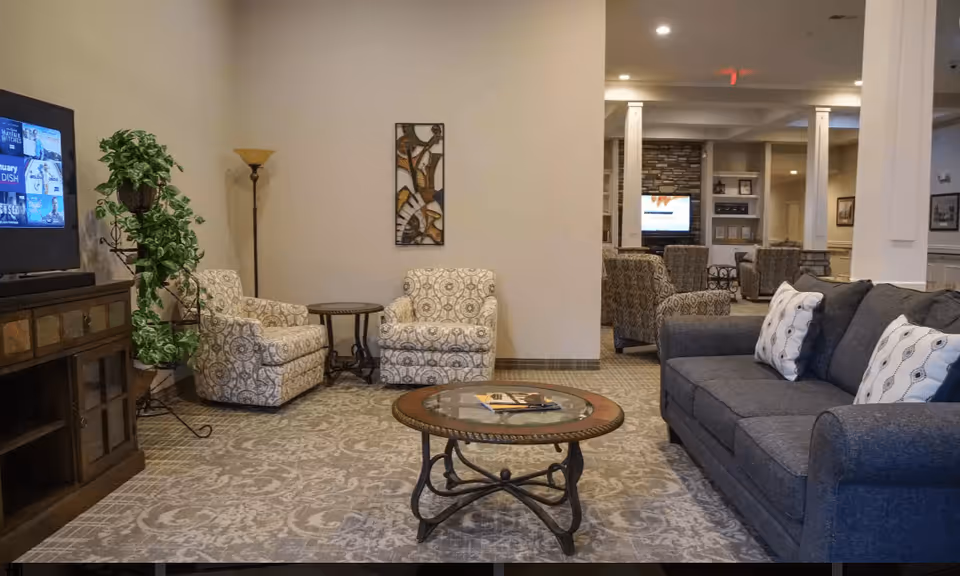 A cozy living room area in a senior living facility featuring a gray sofa with patterned pillows, two patterned armchairs with a small round table between them, a round glass-top coffee table, a tall floor lamp, a potted plant on a stand, and a TV on a wooden cabinet. The room has neutral-colored walls and carpeted flooring with a decorative pattern. In the background, there are more seating areas and a TV mounted on a stone wall.