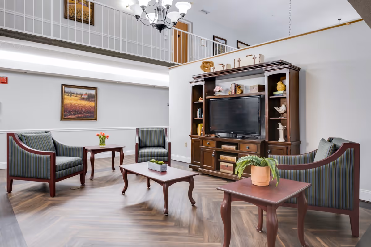 A bright and spacious living room area in a senior living facility with a wooden entertainment center holding a flat-screen TV. The room features three striped upholstered armchairs, two wooden coffee tables with plants and flowers, a chandelier hanging from the ceiling, and framed artwork on the walls. There is a railing and doors visible on an upper level.
