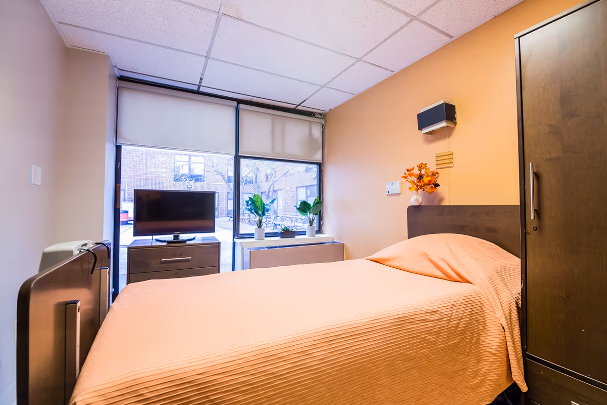 A small, cozy bedroom in a nursing facility with a single bed covered in a peach-colored blanket. There is a dark wooden headboard and a matching wardrobe on the right side. A flat-screen TV sits on a small dresser in front of a large window with partially drawn blinds. Two potted plants and a vase with orange flowers are placed on a white table beneath the window. The walls are painted a warm beige color.