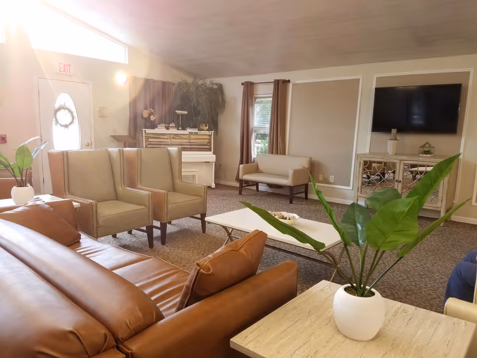 A well-lit living room with a brown leather sofa, two beige armchairs, a beige loveseat, a white coffee table, and a TV mounted on the wall above a decorative cabinet. There are green plants in white pots on tables, a white piano against the wall, and windows with brown curtains letting in natural light.