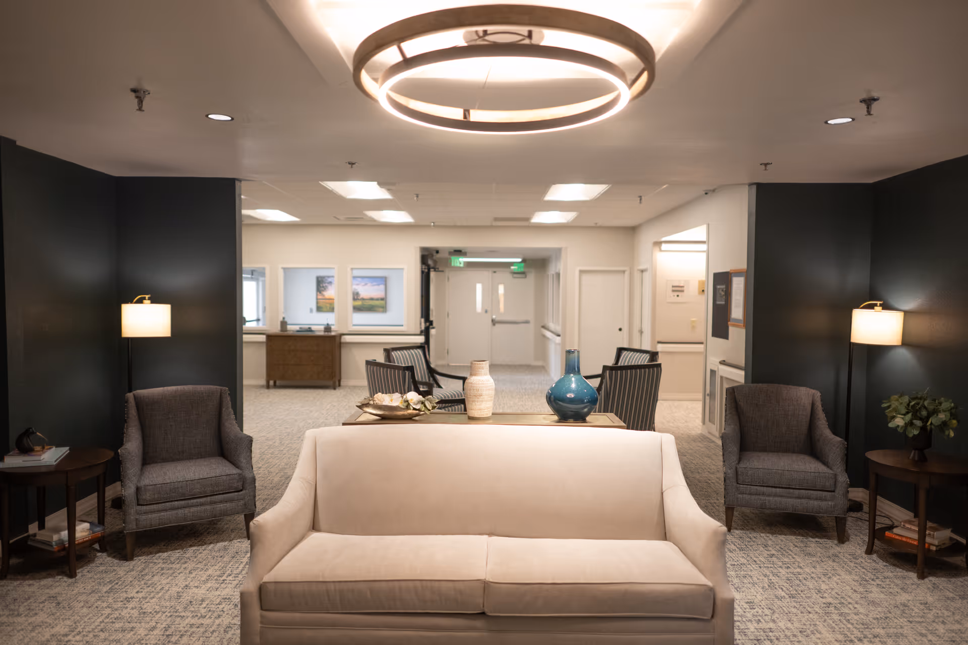 A well-lit senior living facility seating area with a beige sofa, two gray armchairs, side tables, and decorative vases under a circular ceiling light.