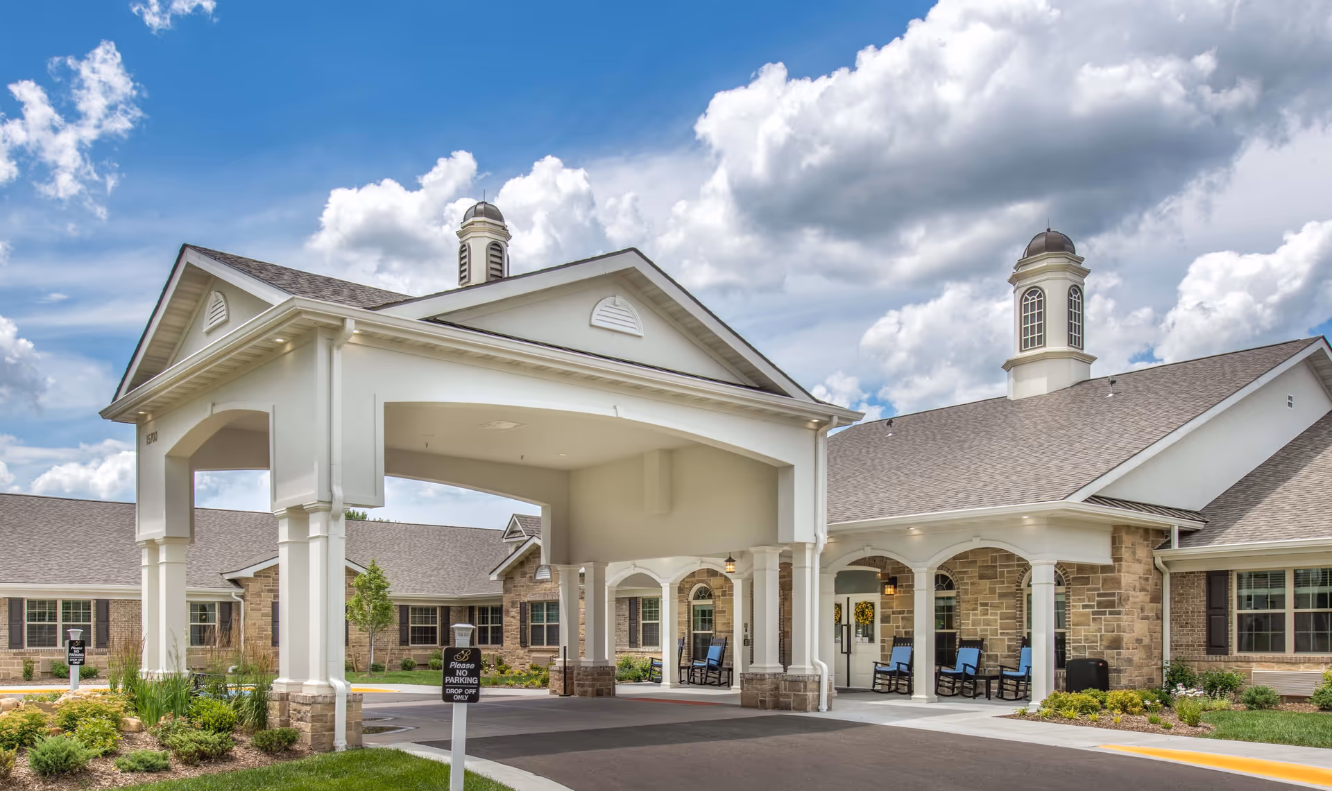 Exterior view of Benton House of Olathe, showing a covered entrance with white pillars, stone and brick facade, multiple windows, and a clear blue sky with clouds above.