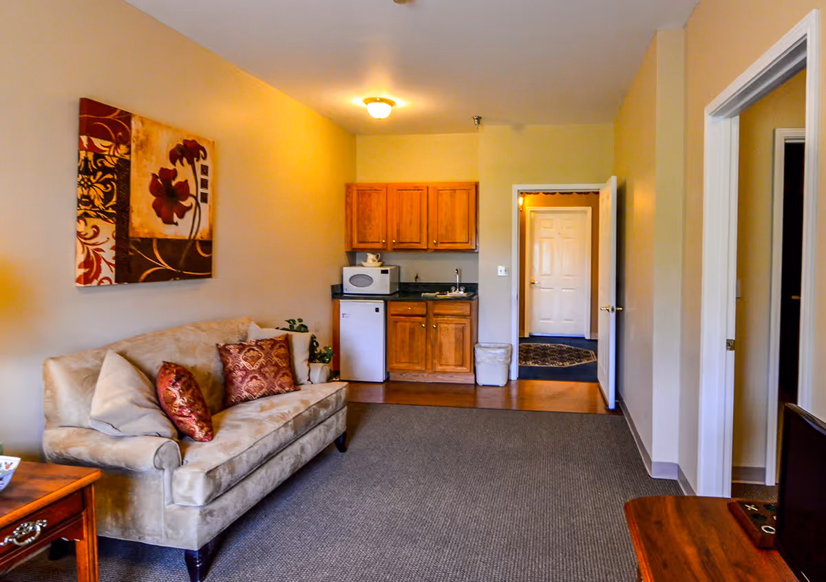 Interior view of a living area in a senior living facility featuring a beige couch with decorative pillows, a small kitchenette with wooden cabinets, a microwave, and a mini fridge. The room has beige walls, a carpeted floor, and a doorway leading to another room.