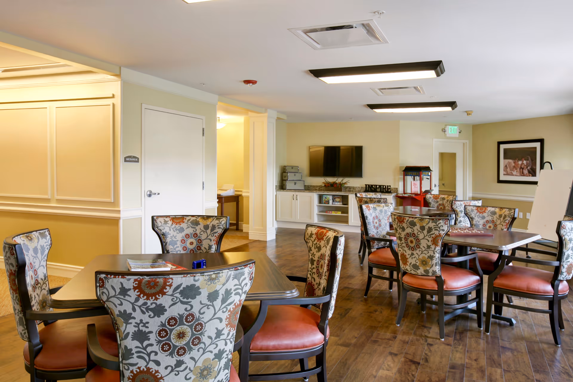 Well-lit communal dining/activity room with patterned upholstered chairs around wooden tables and a TV and cabinets along the far wall.