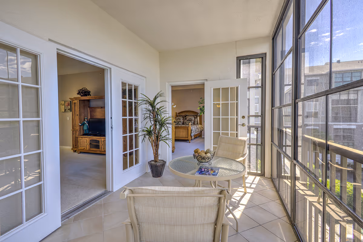 A bright enclosed balcony area with large windows on the right side, two beige chairs, and a round glass table with a decorative bowl and a magazine on it. The balcony leads to two rooms through white French doors, one room with a wooden entertainment center and TV, and the other with a wooden bed and plants.