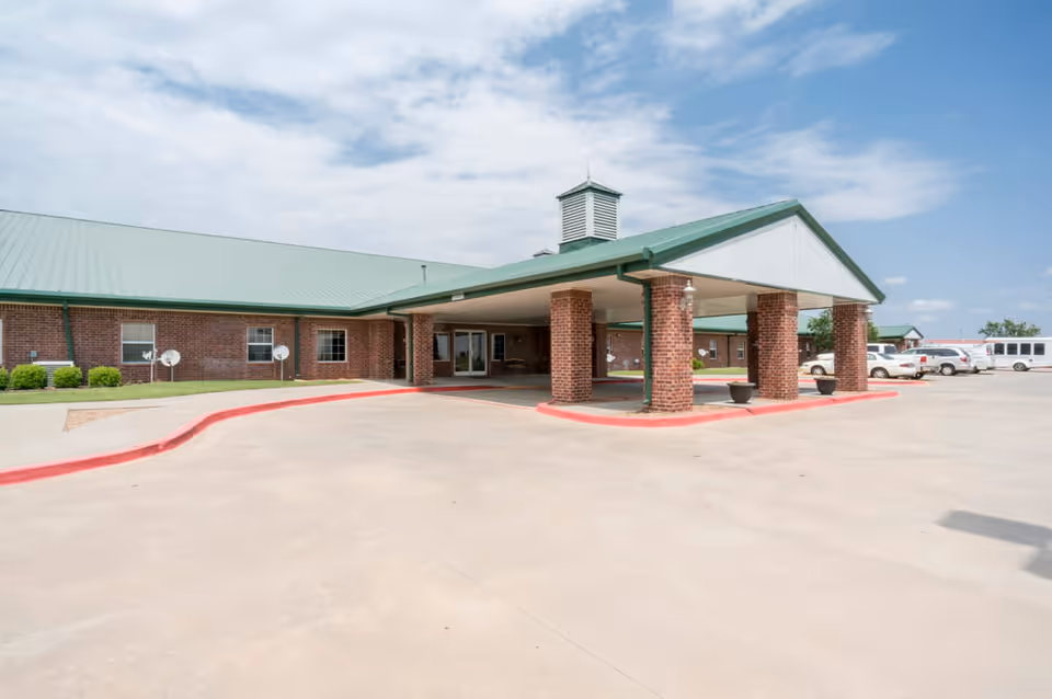 Exterior view of a single-story brick building with a green metal roof and a covered entrance supported by brick columns. Several vehicles are parked nearby under a partly cloudy sky.