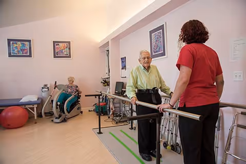 An elderly man using parallel bars for physical therapy assisted by a caregiver in a red shirt, while an elderly woman exercises on a stationary bike in a rehabilitation room with light pink walls and framed artwork.