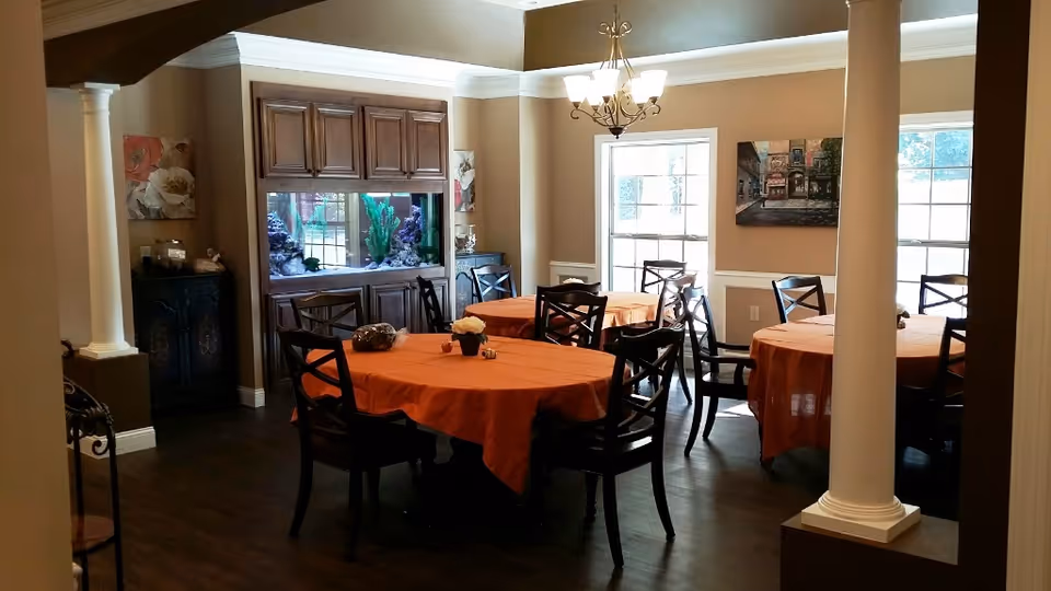 A dining room with three round tables covered with orange tablecloths, each surrounded by dark wooden chairs. The room features two large windows letting in natural light, a chandelier hanging from the ceiling, decorative columns, wall art, and a built-in aquarium with wooden cabinetry.