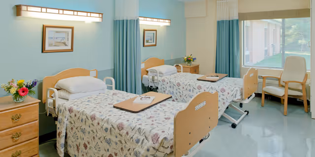 A bright and clean shared bedroom in a senior living facility with two hospital-style beds covered with floral bedspreads. Each bed has a wooden headboard and a tray table. There is a wooden nightstand with a vase of colorful flowers between the beds. A large window with blinds lets in natural light, and a comfortable armchair is placed near the window. Light blue and white curtains hang between the beds for privacy.