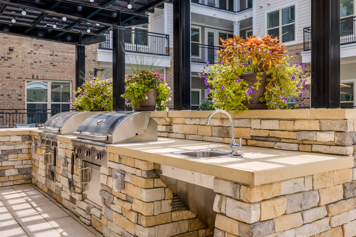 Outdoor kitchen area with two stainless steel grills and a sink set in a stone countertop. There are potted plants with colorful flowers placed on the countertop. The background shows a multi-story building with balconies and windows.