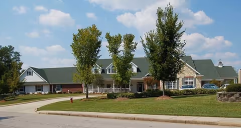 Front exterior of a single-story senior living building with a covered porch, green roof, trees, and landscaped lawn.