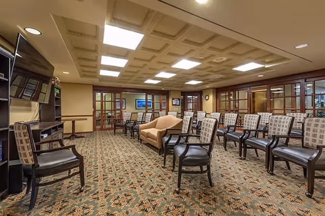 A spacious common area with patterned carpet, multiple rows of chairs arranged facing a wall-mounted television, a beige couch in the center, and wooden framed glass doors along the back wall. The ceiling has recessed lighting panels.