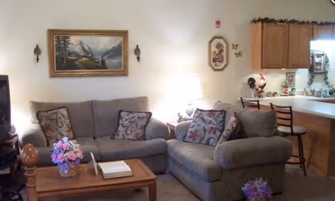 Living room with a sofa and armchair, coffee table, lamp, wall art, and an open kitchen counter with bar stools.