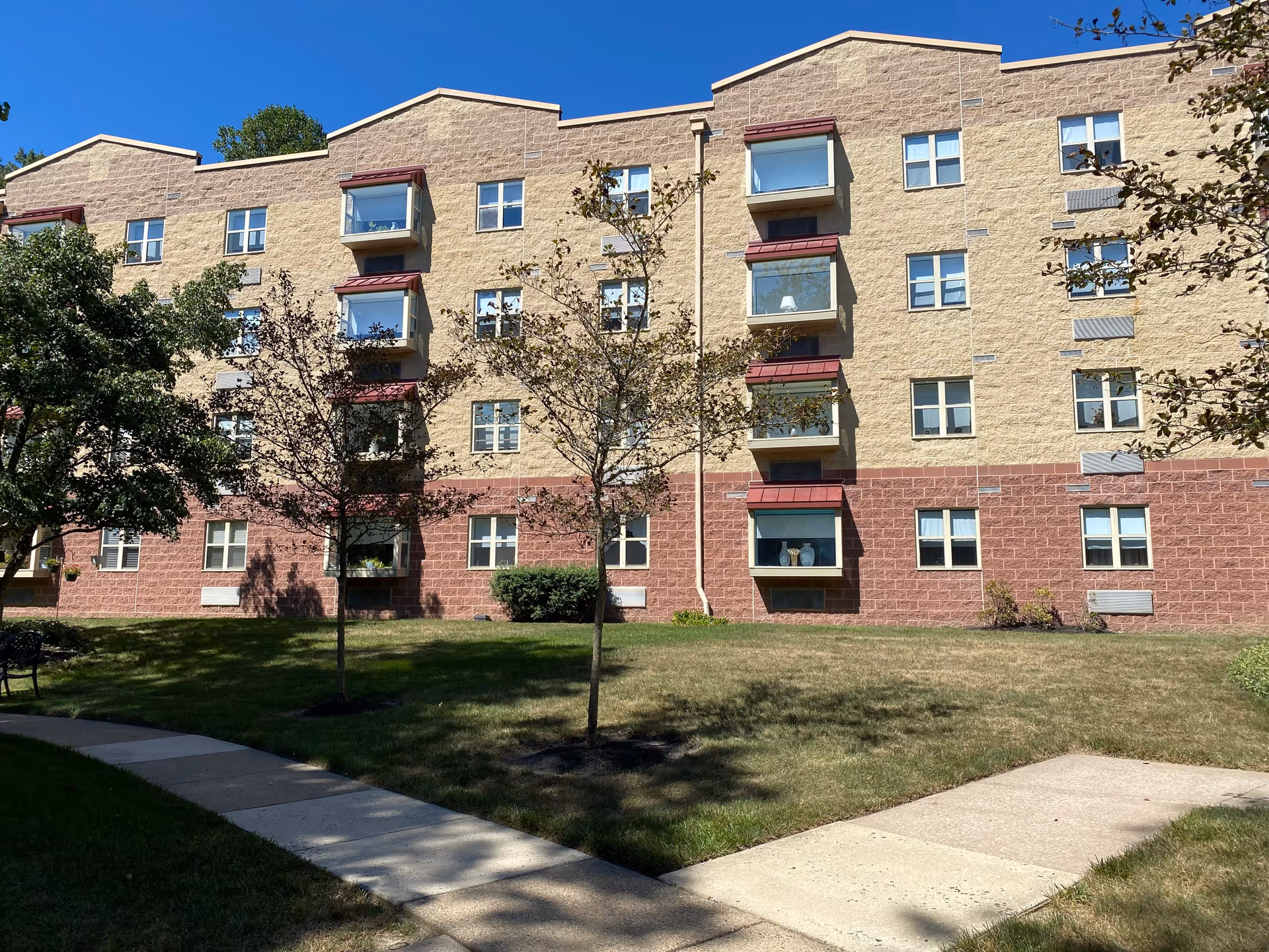 Exterior view of a multi-story senior living facility building with beige and reddish-brown brick walls, several windows, and small balconies. There are a few young trees and a grassy lawn in front of the building, along with a concrete walkway. The sky is clear and blue.