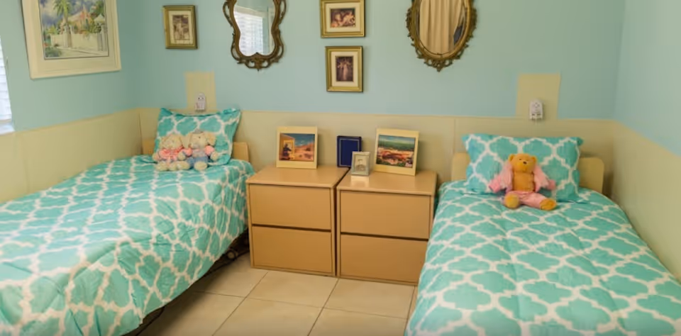 A bedroom with two single beds featuring turquoise and white patterned bedding. Each bed has a stuffed animal on the pillow. Between the beds are two beige nightstands with framed pictures and a small clock. The walls are painted light blue and beige, decorated with two ornate mirrors and several framed pictures.