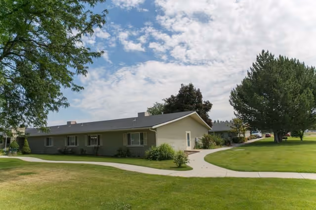 Single-story ranch-style senior living building with a green lawn, trees, and curved walkways under a partly cloudy sky.