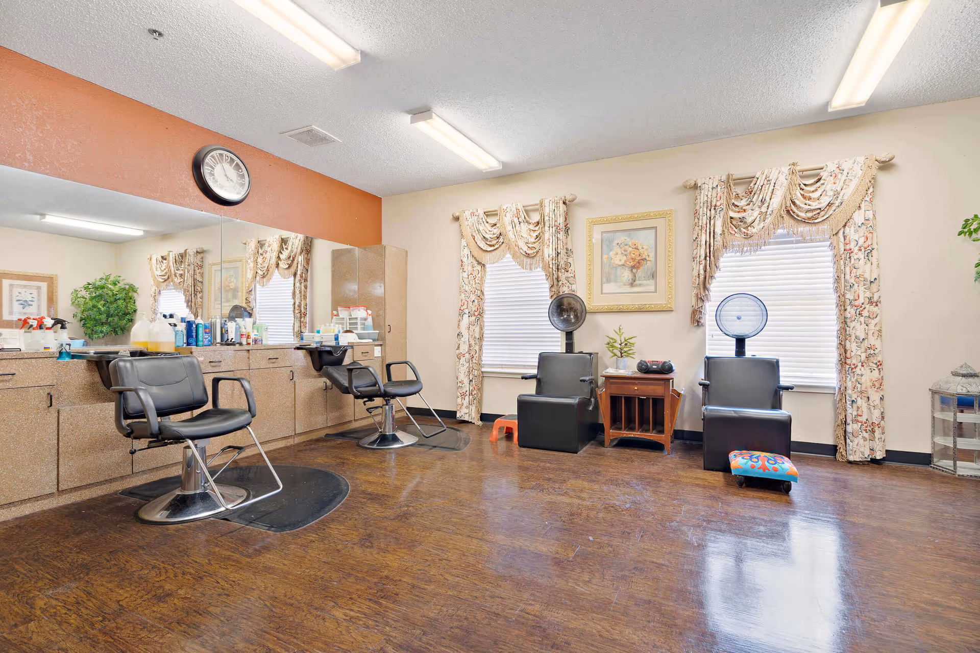 Interior of a salon room with two black salon chairs in front of a large mirror and counter with hair products. Two black hair dryer chairs are positioned near two windows with floral curtains. A small wooden table with a plant and a radio is between the hair dryer chairs. The floor is wooden and the walls are painted beige with one accent wall in orange.