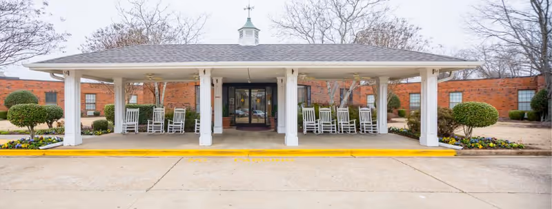 Front entrance of a single-story brick building with a covered porch supported by white columns. Several white rocking chairs are arranged under the porch. There are neatly trimmed bushes and flower beds on either side of the entrance. Leafless trees are visible in the background.