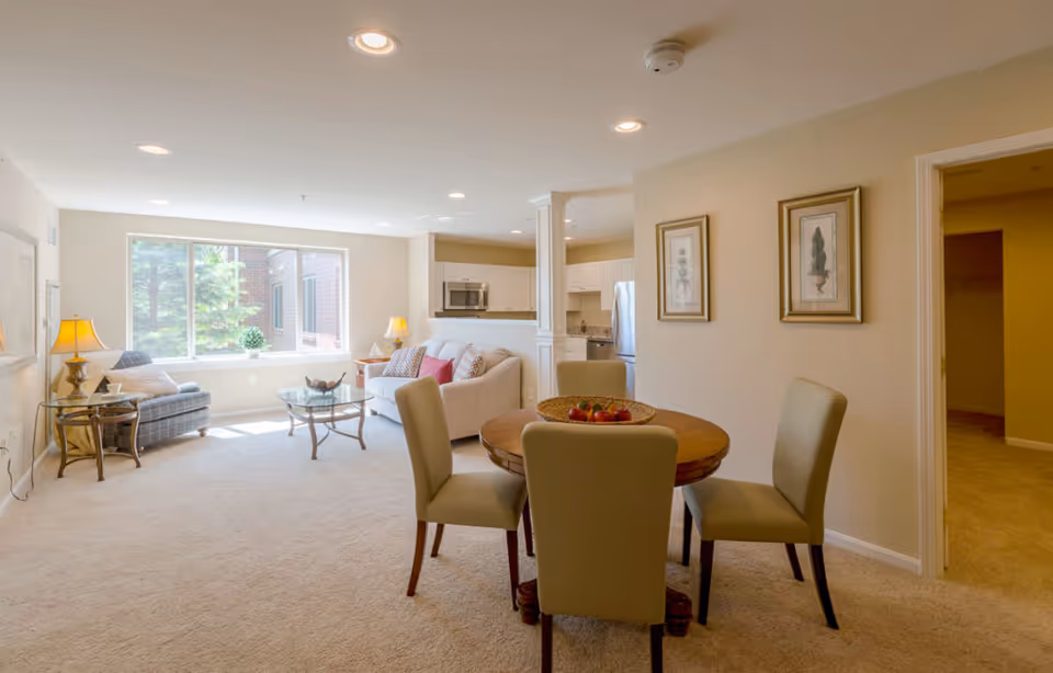 Bright open living and dining area with a round wooden table and chairs, sofa and armchair near a large window, and a view into the kitchen.