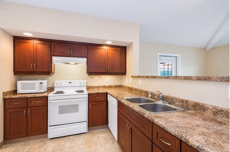 A kitchen with wooden cabinets, a white electric stove with an oven, a white microwave on the countertop, a double stainless steel sink, and a dishwasher. The countertops are a brown granite pattern, and the floor is tiled. There is a small window above the sink area.