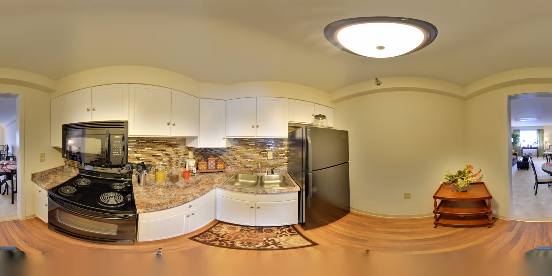A compact kitchen with white cabinets, a black stove and microwave, a double sink, and a black refrigerator. The countertop has various kitchen items including jars and a coffee maker. The backsplash features a mosaic tile design. There is a small wooden table with a floral arrangement on the right side. Two doorways lead to other rooms, one showing a dining area and the other a living room.