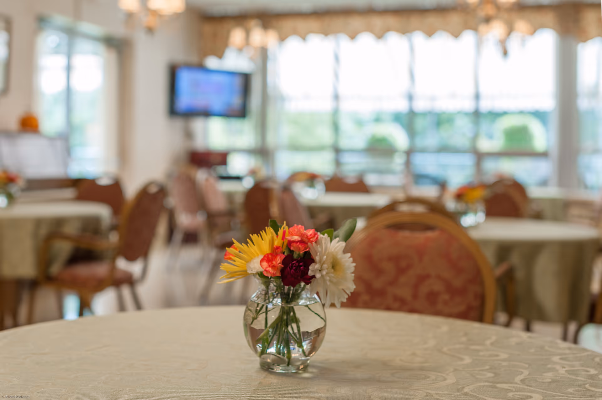 A dining room with round tables covered in beige tablecloths, each table decorated with a small glass vase holding colorful flowers. Chairs with patterned upholstery surround the tables. Large windows in the background let in natural light, and a television is mounted on the wall.