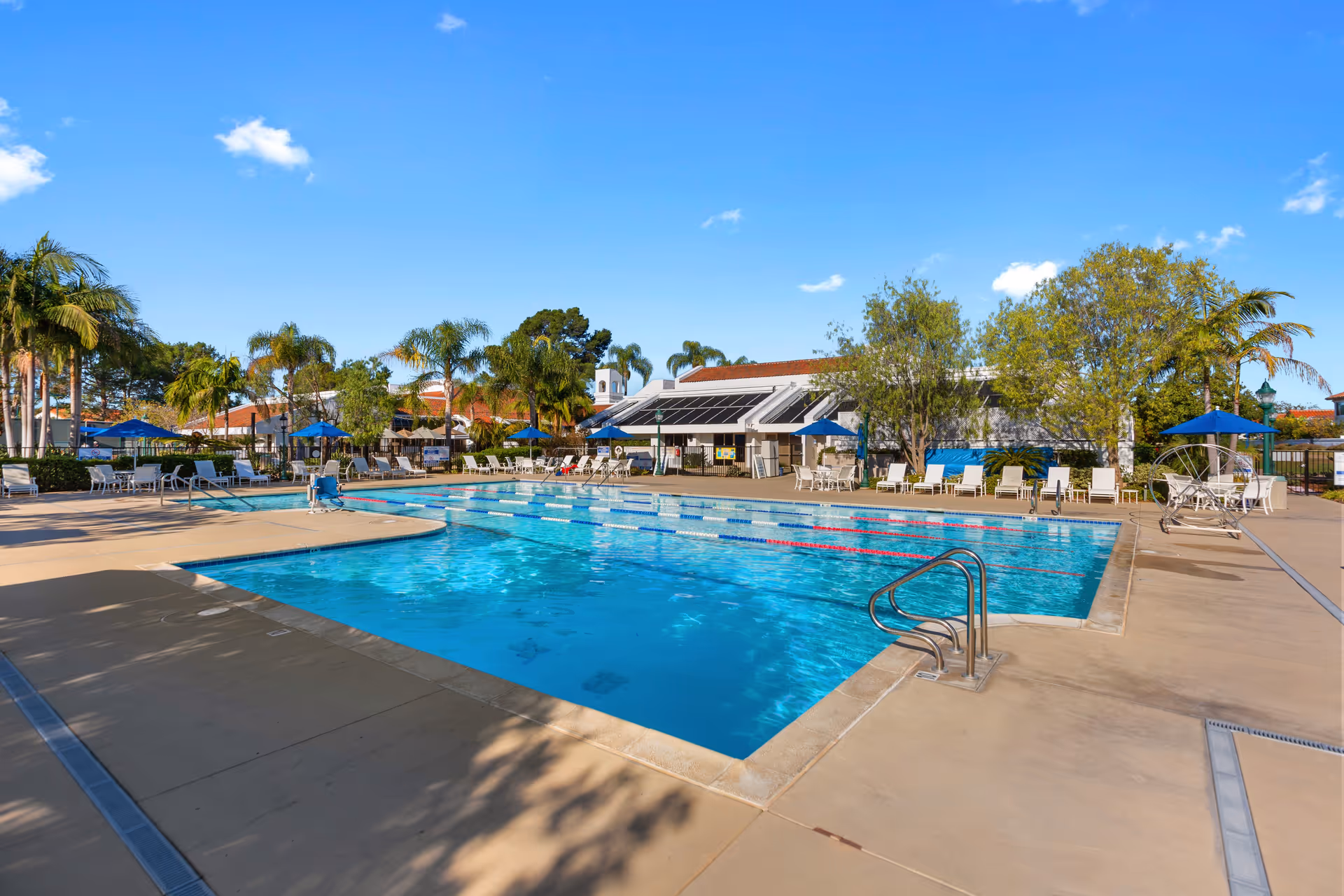 Outdoor swimming pool with clear blue water, surrounded by lounge chairs and tables with blue umbrellas. Palm trees and other greenery are visible around the pool area, with a building in the background under a clear blue sky.