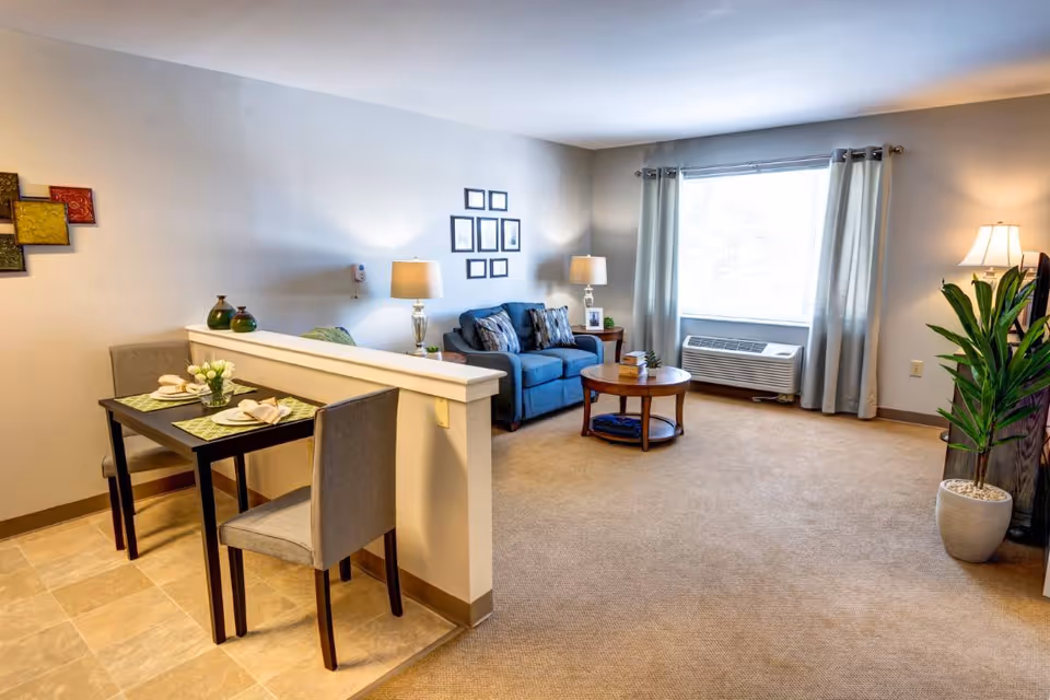 Bright furnished living room with a small dining nook, sofa, coffee table, side lamps, and a large window with curtains.