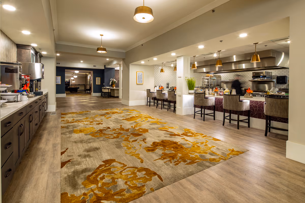 Interior view of a senior living facility dining and kitchen area with a large patterned rug on the wooden floor, a long counter with chairs, pendant lights hanging from the ceiling, and a kitchen area with stainless steel appliances and white subway tile backsplash.