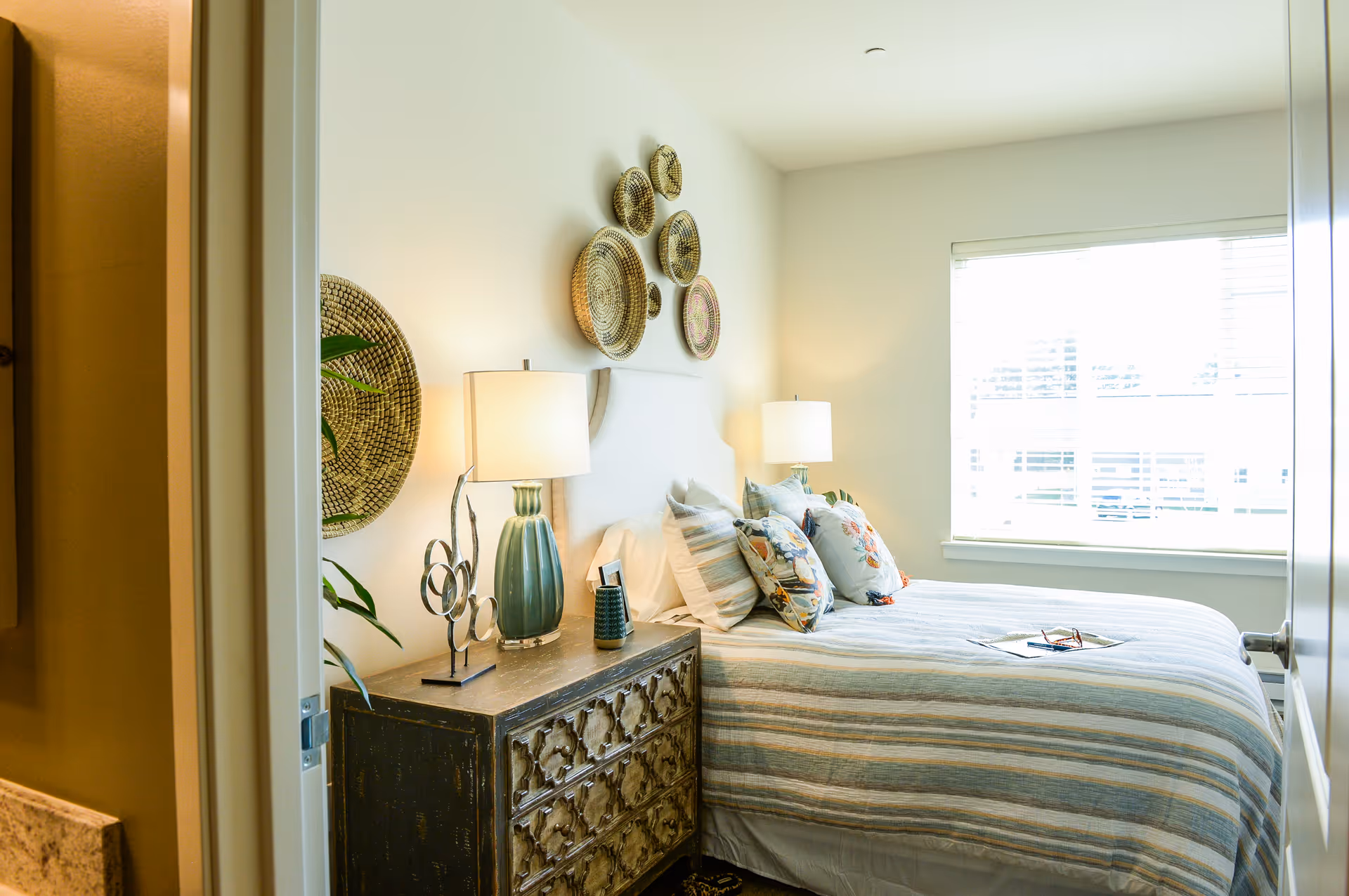 Bright bedroom with a striped bed, decorative pillows, a nightstand with a lamp and decor, and woven wall baskets above the headboard.