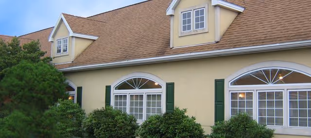Cream-colored building facade with arched windows, green shutters, dormer windows, and bushes in front.
