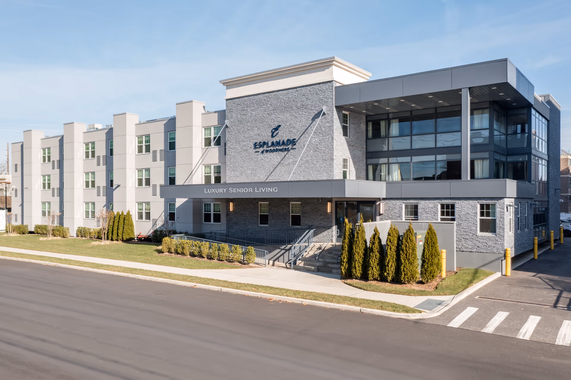 Exterior view of Esplanade of Woodmere, a modern three-story senior living facility with a combination of gray stone and light-colored walls, large windows, and a covered entrance with a ramp and stairs. The building is surrounded by neatly trimmed bushes and a well-maintained lawn under a clear blue sky.