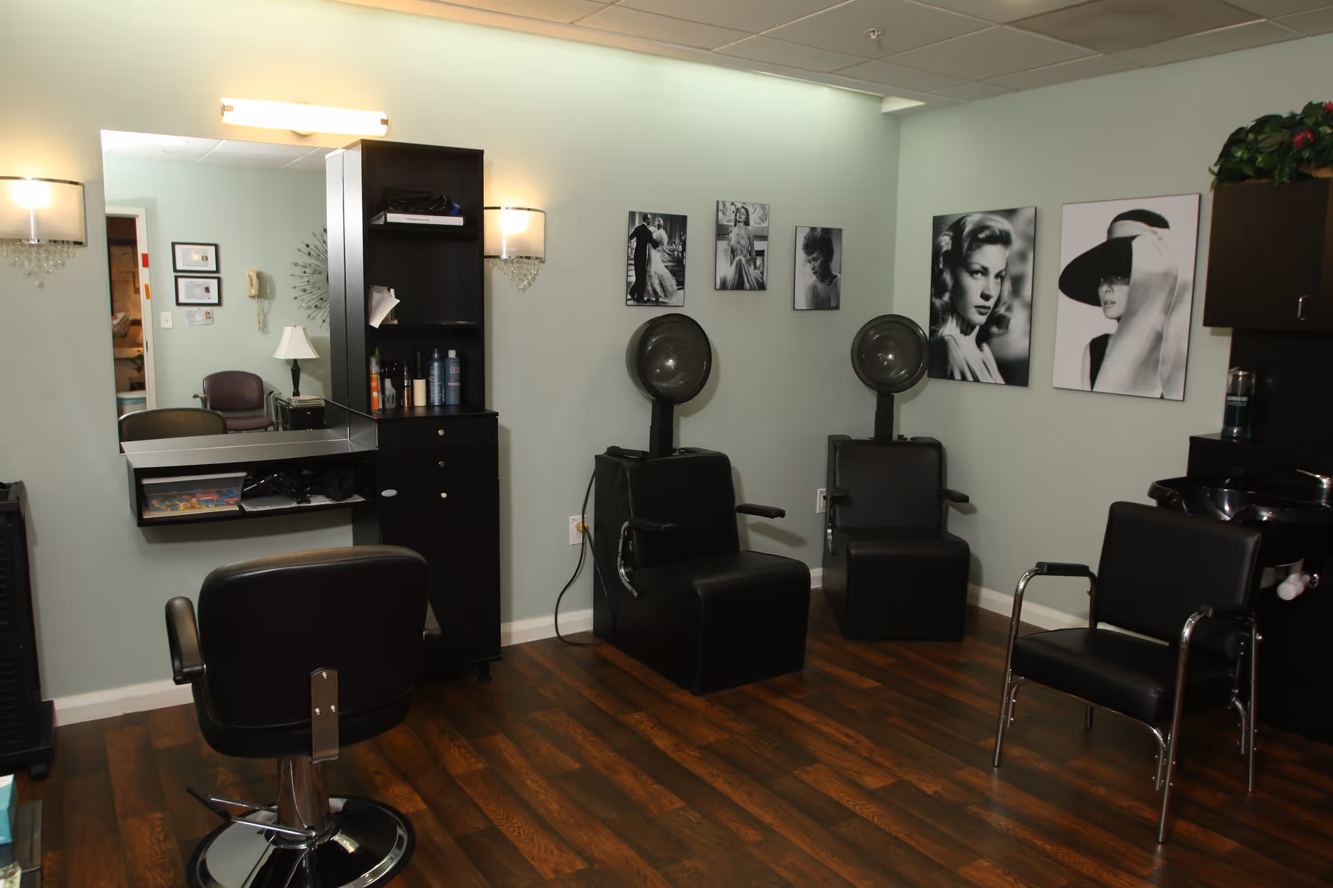 Interior of a hair salon area with three black salon chairs, two hair dryers, a large mirror with a shelf holding hair products, and black and white portraits of women on the walls. The floor is wooden and the walls are painted light green.