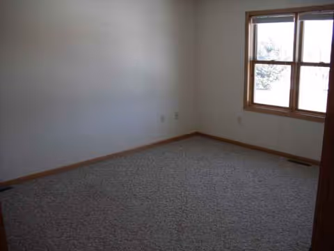Empty carpeted room with a window, bare walls, and wooden trim.