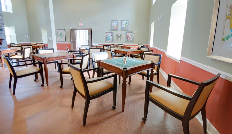 A bright and spacious common area with multiple wooden tables and cushioned chairs arranged for group activities or socializing. The room has large windows letting in natural light, light green walls with a red lower border, framed artwork, and a door at the far end.