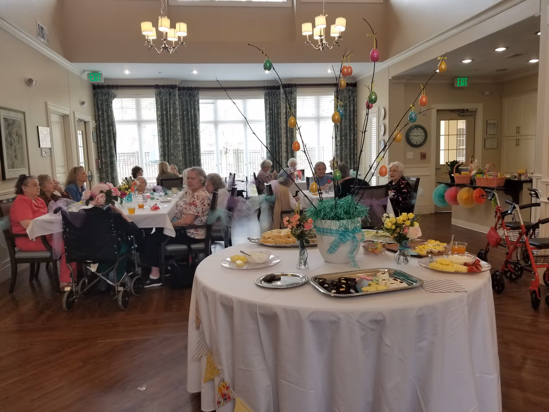 A group of elderly people seated around tables in a well-lit dining room with large windows and green curtains. In the foreground, a round table is decorated with a white tablecloth, a centerpiece with branches and colorful hanging ornaments, and trays of cookies and snacks. The room has wooden floors, chandeliers, and exit signs above doors.