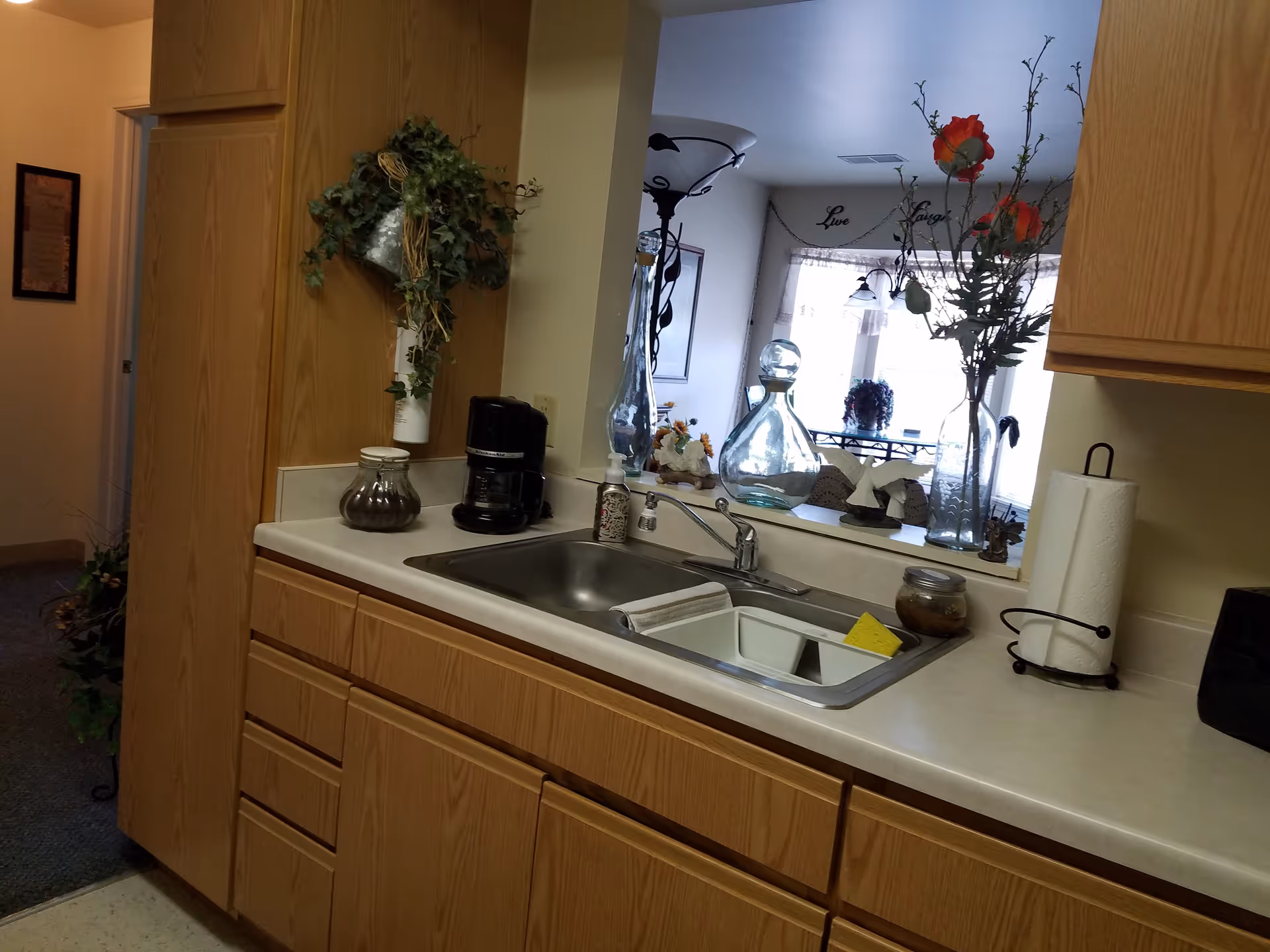 Interior view of a kitchen area with wooden cabinets, a stainless steel sink with a sponge and soap dispenser, a coffee maker, a paper towel holder, and decorative plants and glass vases on the counter and window ledge. Through the window opening, a dining area with a table and chairs is visible.