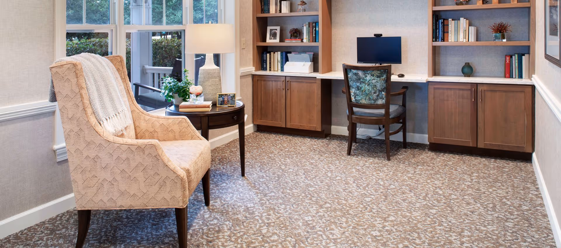 A cozy interior room with a patterned beige armchair draped with a light throw blanket next to a round wooden side table holding a lamp, a small plant, books, and a framed photo. In the background, there is a built-in wooden desk with a computer monitor and a chair with a floral backrest, flanked by bookshelves filled with books and decorative items. Large windows let in natural light and show greenery outside.