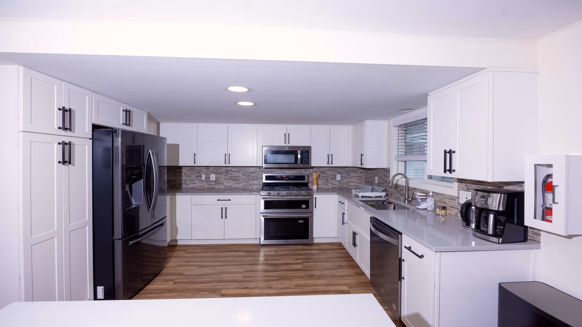 A modern kitchen with white cabinets, stainless steel appliances including a refrigerator, oven, microwave, and dishwasher. The kitchen has a light gray countertop, a coffee maker, a sink under a window with blinds, and a fire extinguisher mounted on the wall. The floor is wood-style laminate.