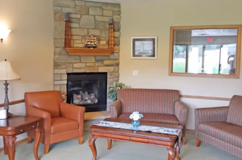 A cozy living room area with a stone fireplace in the center. There is a brown leather armchair to the left, a patterned loveseat in the middle, and a matching armchair to the right. A wooden coffee table with a decorative runner and a small vase of flowers sits in front of the loveseat. A side table with a lamp is next to the leather armchair. A framed picture hangs on the wall above the loveseat, and a large window reflects the outside view.