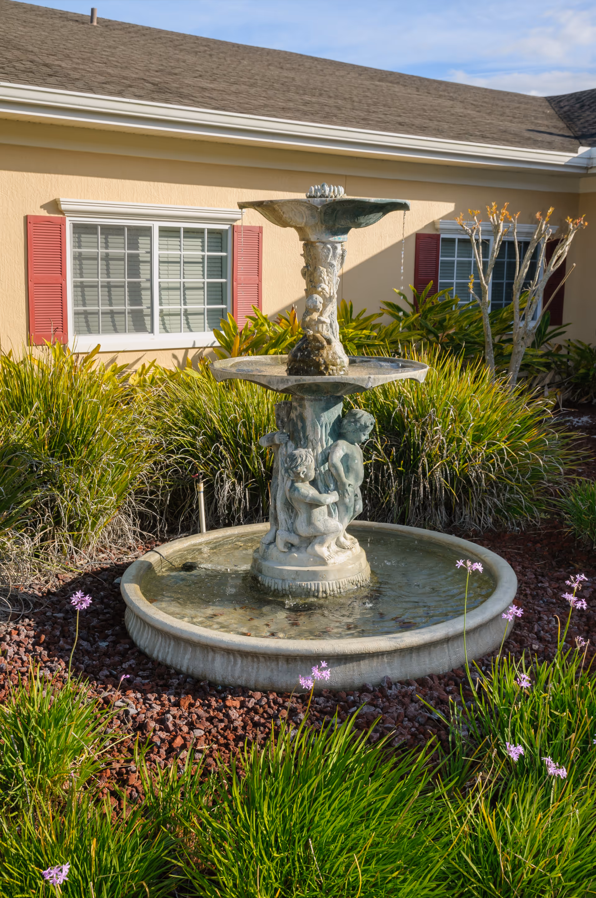 Outdoor stone fountain with sculptures of children, surrounded by green plants and purple flowers, in front of a beige building with red window shutters.