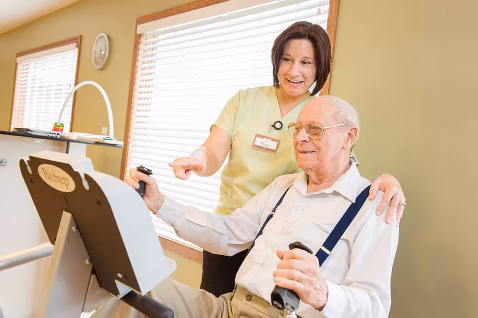 A senior man wearing glasses and suspenders is using a seated exercise machine while a female caregiver or therapist in a light green uniform stands beside him, smiling and pointing at the machine's controls in a well-lit room with large windows and beige walls.