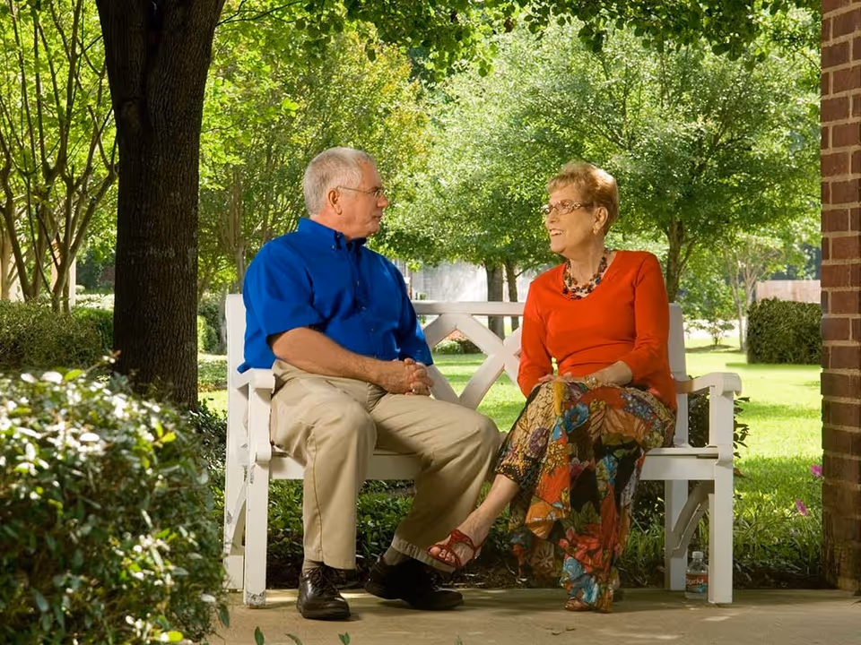 An elderly man and woman sitting on a white wooden bench outdoors, engaged in conversation. The man is wearing a blue shirt and beige pants, while the woman is dressed in a red top and colorful floral skirt. They are surrounded by green trees and bushes, with sunlight filtering through the leaves.