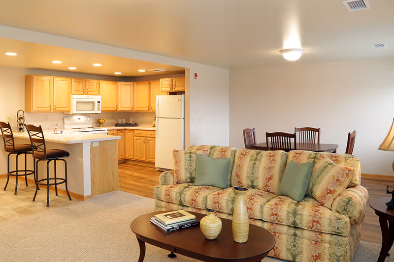 A cozy living room and kitchen area in a senior living facility. The living room features a floral patterned sofa with green cushions, a wooden coffee table with decorative vases and books, and a side table with a lamp. The kitchen has light wood cabinets, a white refrigerator, microwave, stove, and a breakfast bar with two high chairs. In the background, there is a dining area with a wooden table and four chairs.