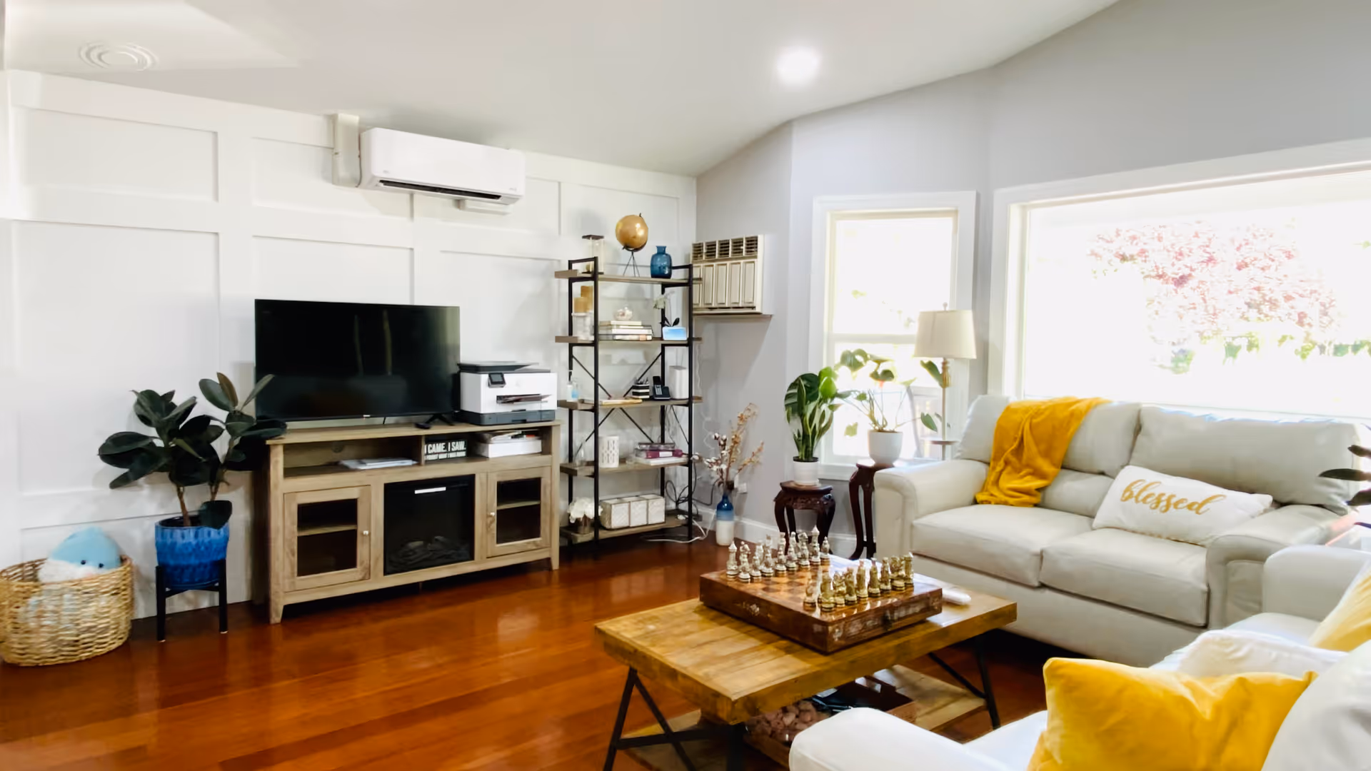 A bright and cozy living room with wooden flooring, a white couch with yellow and white pillows, a wooden coffee table with a chess set, a TV on a wooden stand, a black metal shelf with decorative items, and several potted plants near large windows letting in natural light.